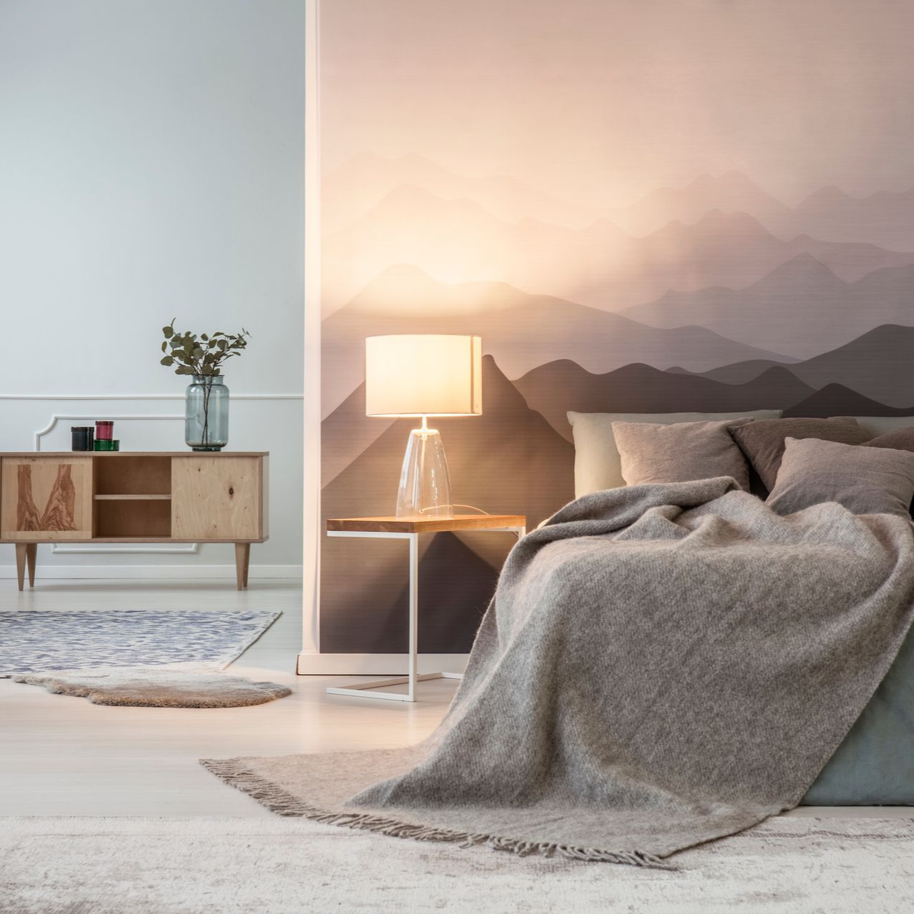 Chambre moderne avec lit, lampe, armoire en bois et linge de lit gris clair sur fond de fresque murale représentant un lever de soleil sur une montagne