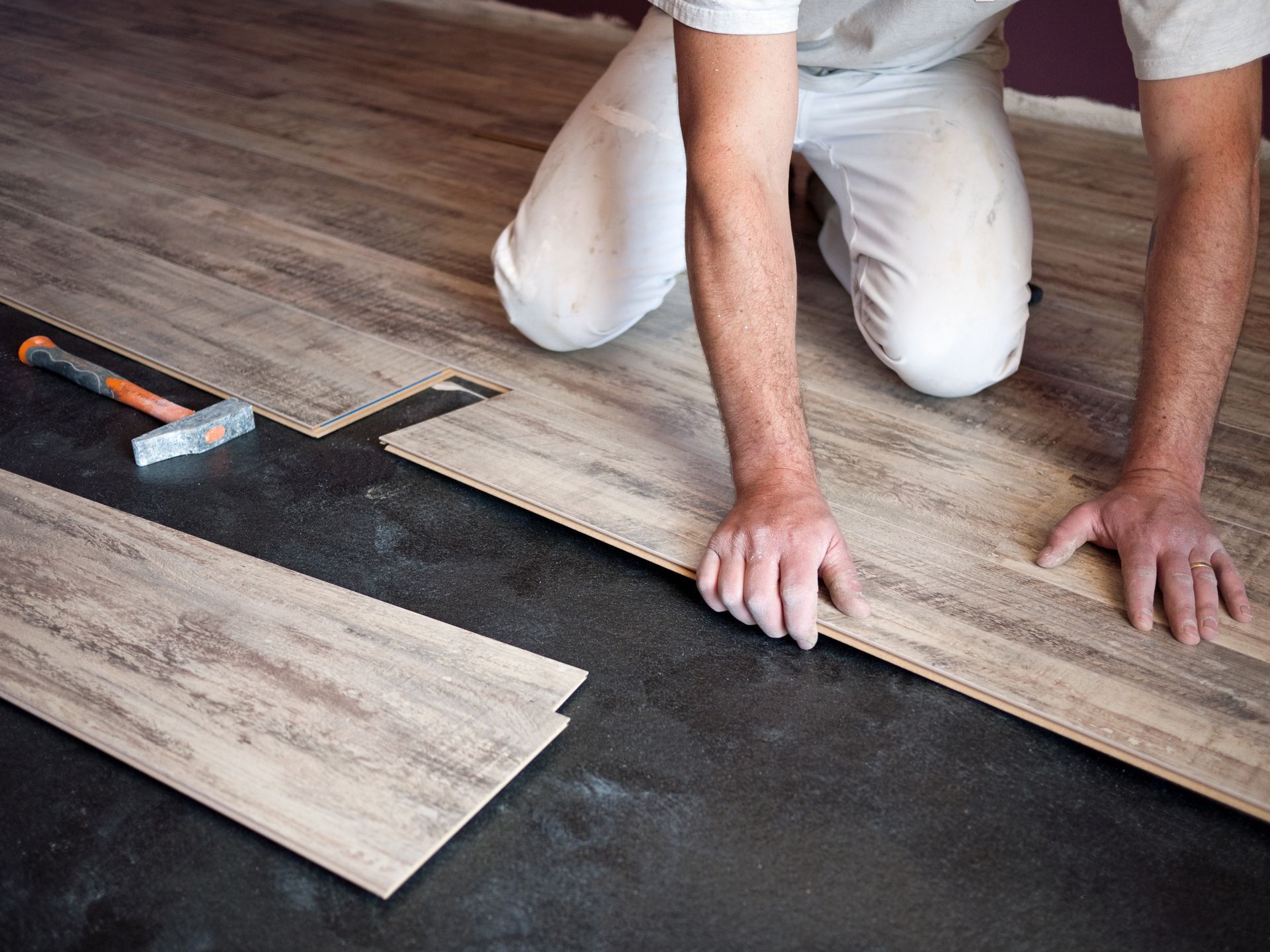 Une personne installe un revêtement de sol imitation en bois à l'aide d'un marteau et de cales d'espacement sur un sous-plancher foncé.