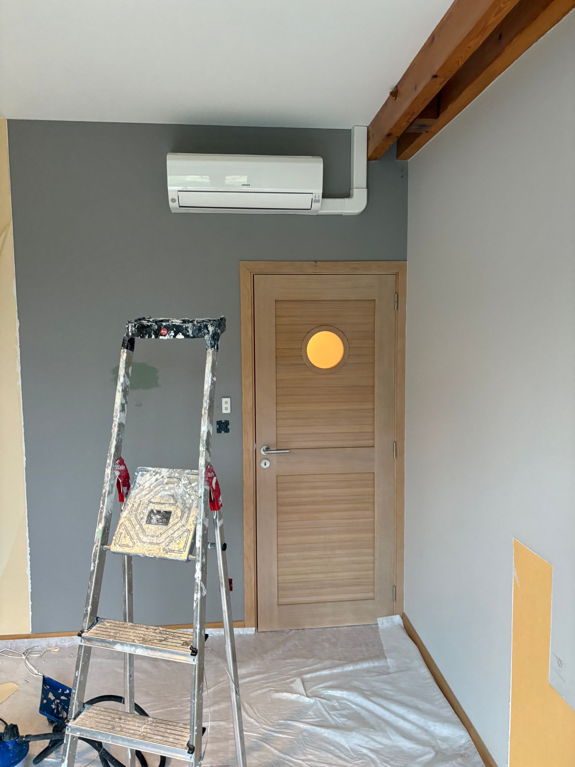 Pièce intérieure en rénovation avec échelle, sol recouvert de plastique, mur gris, porte en bois et climatiseur mural