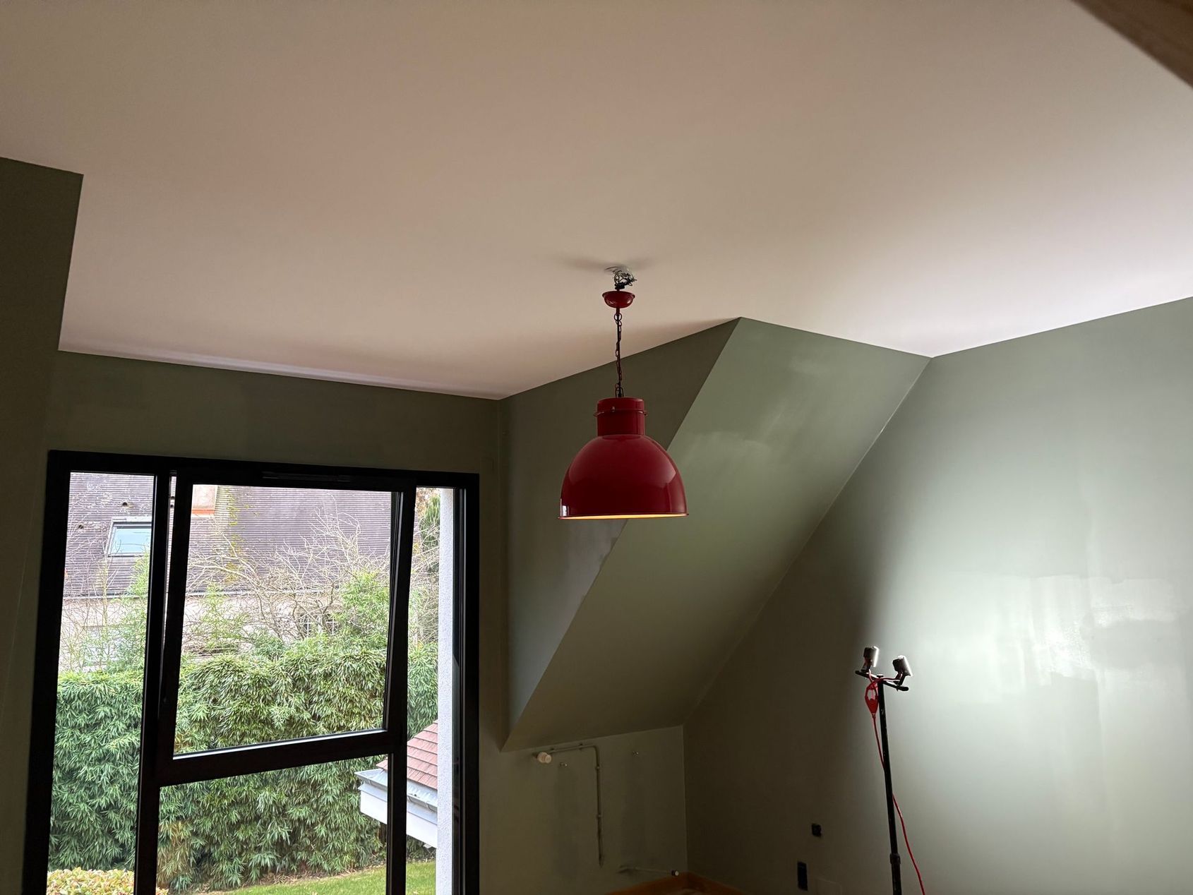 Pièce vide aux murs verts, avec une lampe suspendue rouge et une fenêtre donnant sur des arbres.
