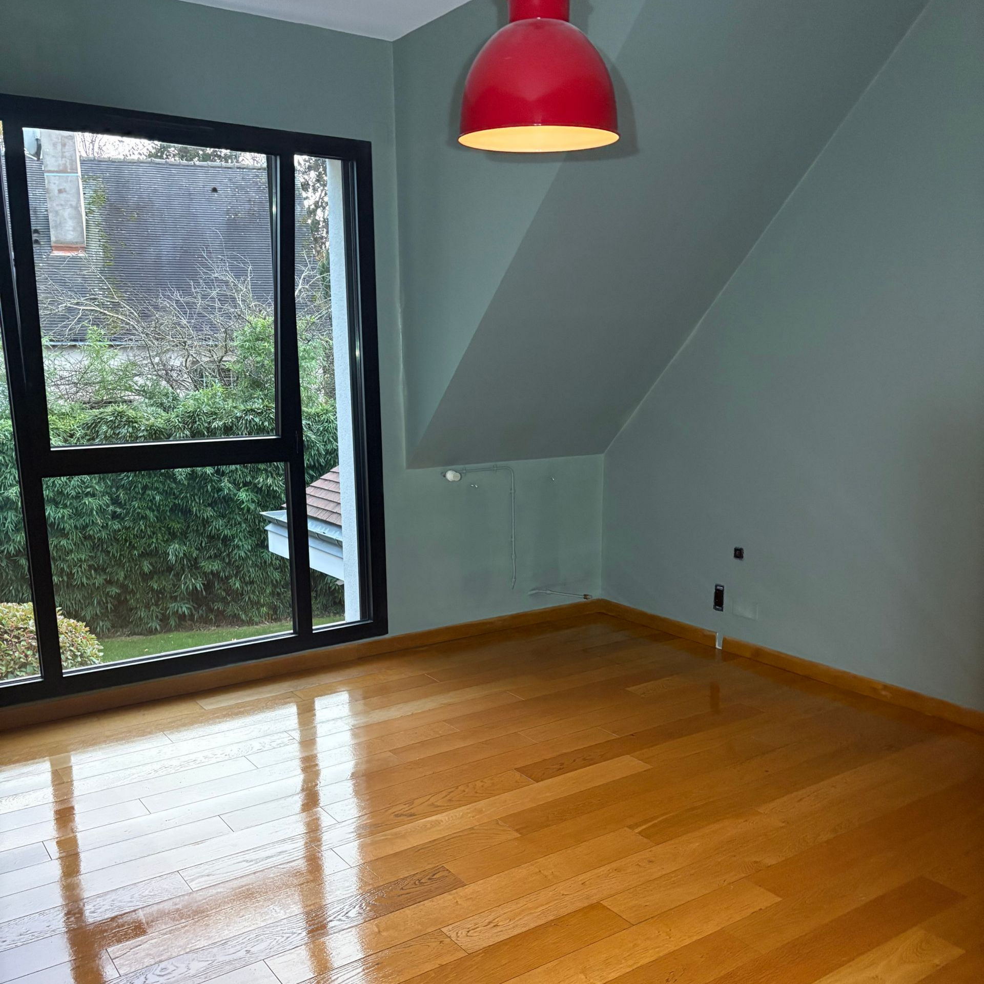 Pièce vide avec parquet, murs gris, grande fenêtre et suspension rouge