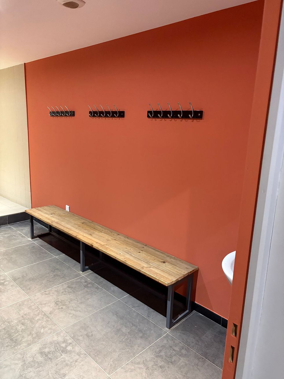 Couloir vide avec un long banc en bois adossé à un mur corail et plusieurs patères noires.