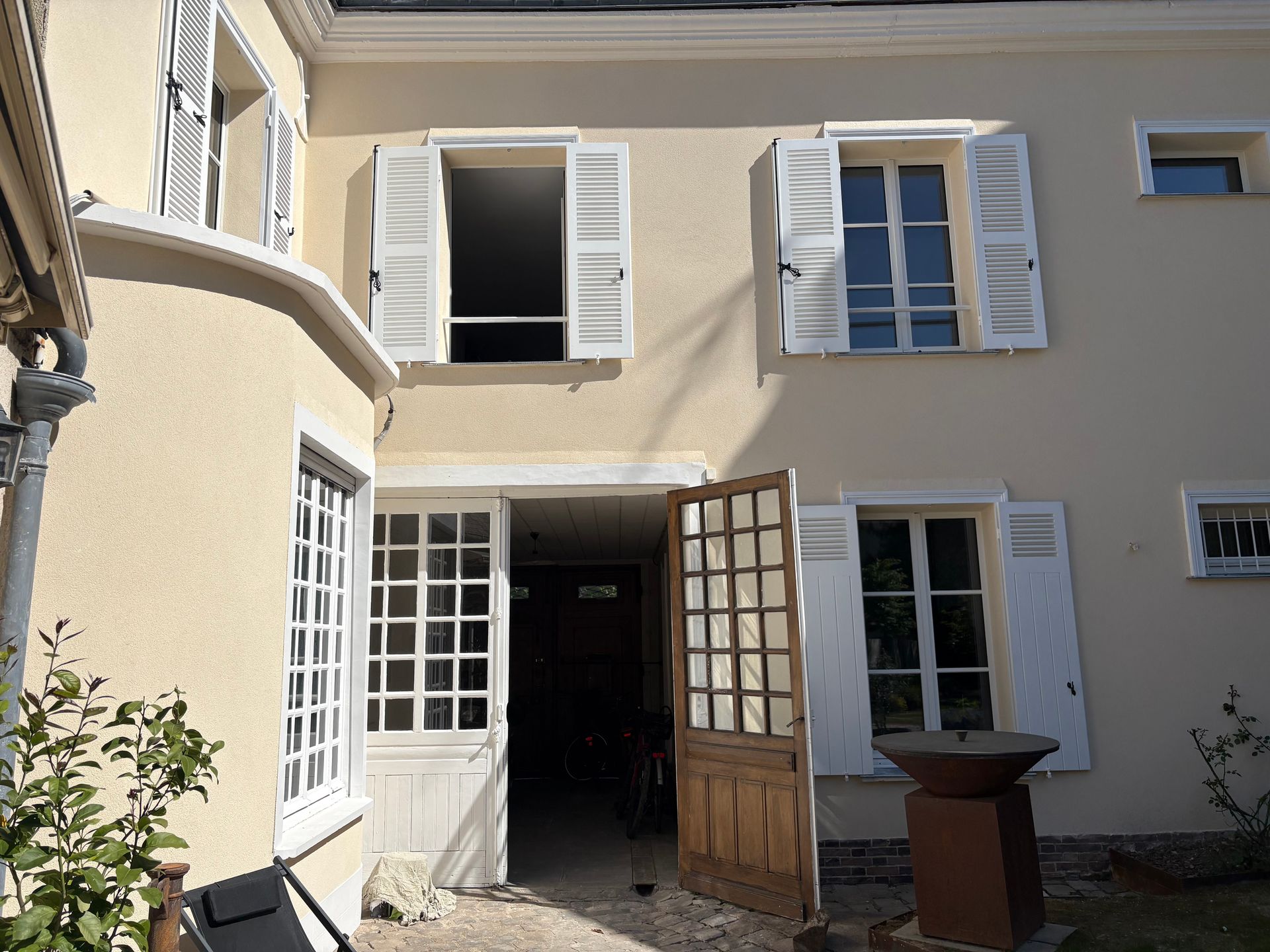 Entrée par une cour intérieure d'un immeuble beige avec des volets blancs ouverts et des portes doubles vitrées