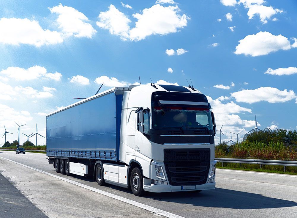 Weißer Sattelschlepper fährt auf einer Autobahn unter blauem Himmel mit Wolken, im Hintergrund Windturbinen.