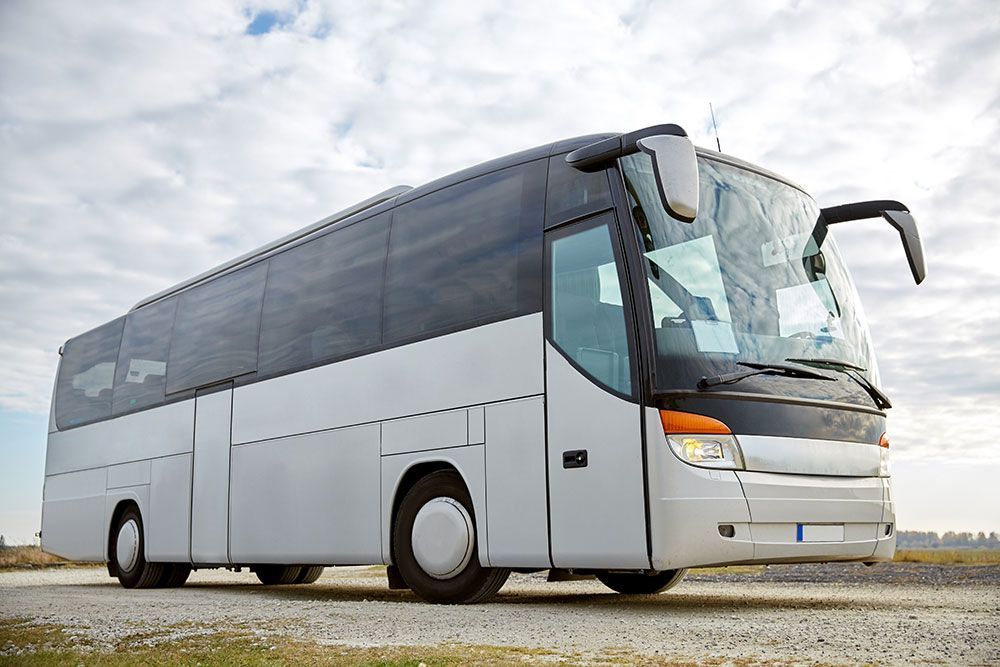 Grau-weißer Personenbus auf einer unbefestigten Straße vor bewölktem Himmel.