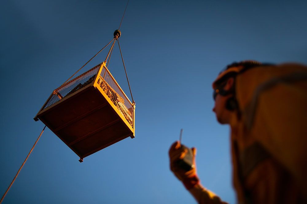 Person, die einen Kran bedient und einen Holzkäfig vor einem blauen Himmel anhebt.