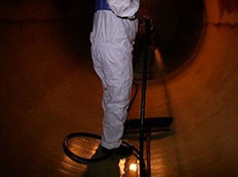 Persona con traje de protección limpiando el interior de un gran contenedor cilíndrico con una manguera.