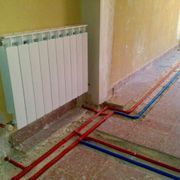 Radiador blanco en la pared, con tubos rojos y azules que recorren el suelo, en una habitación en construcción.