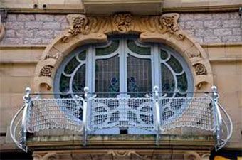 Balcón adornado de estilo Art Nouveau con una ventana arqueada azul y una barandilla de metal decorativa.