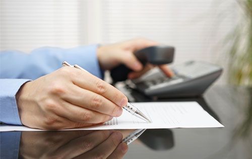Manos escribiendo en papel, sosteniendo el auricular del teléfono, al lado de un teléfono de escritorio negro.