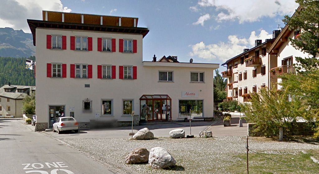 Ein mehrstöckiges beigefarbenes Gebäude mit roten Fensterläden steht neben einem Wohnhaus auf einem gepflasterten Platz mit grossen Steinen.