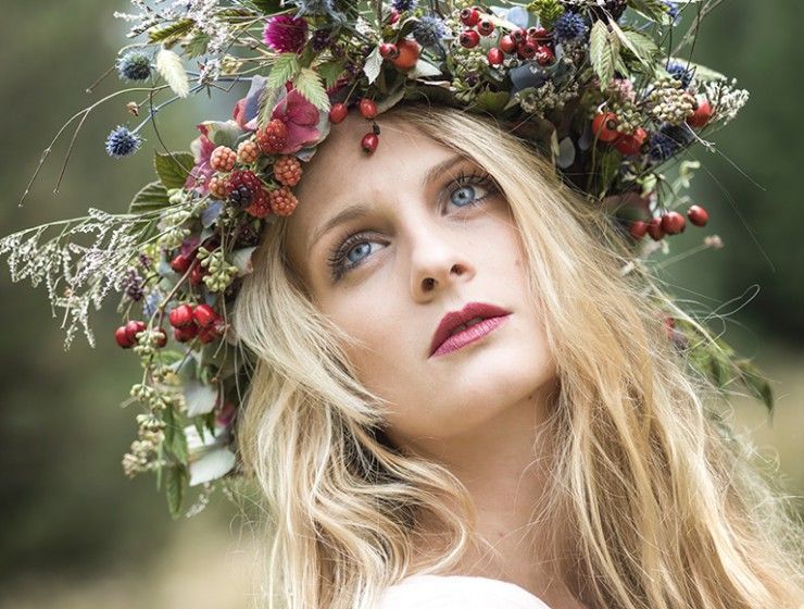 Eine Person mit blonden Haaren trägt einen Kranz aus Beeren, Blumen und Blättern und blickt mit gelassenem Ausdruck nach oben.