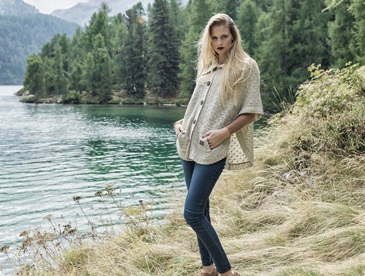 Eine blonde Person steht an einem See, trägt eine beige, kurzärmelige Jacke mit Knöpfen und blaue Jeans und schaut in die Kamera.
