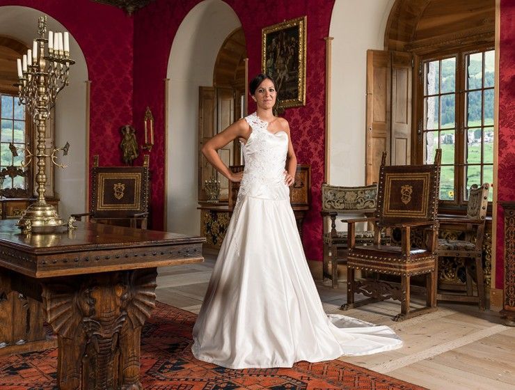 Eine Person in einem weissen Brautkleid steht mit in die Hüften gestemmten Händen in einem reich verzierten Raum mit rot gemusterten Wänden und Holzstühlen.