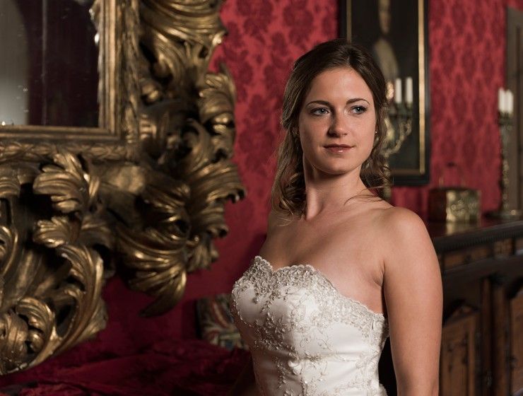 Eine Person in einem weissen, verzierten, trägerlosen Kleid posiert vor einer rot gemusterten Wand neben einem grossen, reich verzierten Goldspiegel.