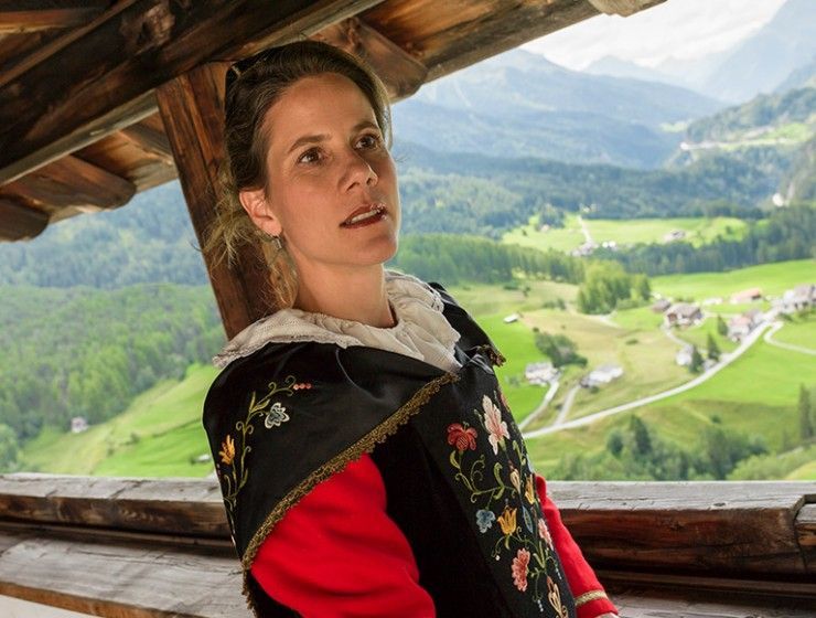 Eine Person in traditioneller, bestickter Tracht blickt von einem Holzbalkon aus auf ein grünes Bergtal.