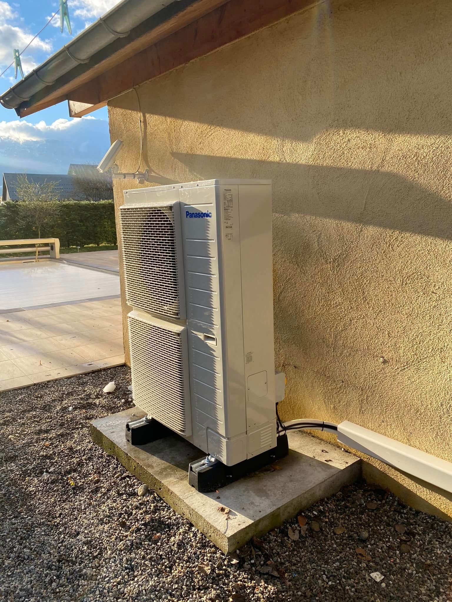 Unité de climatisation extérieure adossée à un mur beige d'une maison sur une dalle de béton, avec un sol en gravier et un ciel bleu.