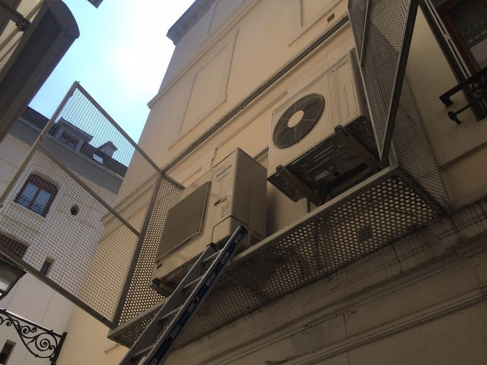 Deux unités de climatisation fixées au mur d'un bâtiment, une échelle appuyée contre elles dans une ruelle étroite.