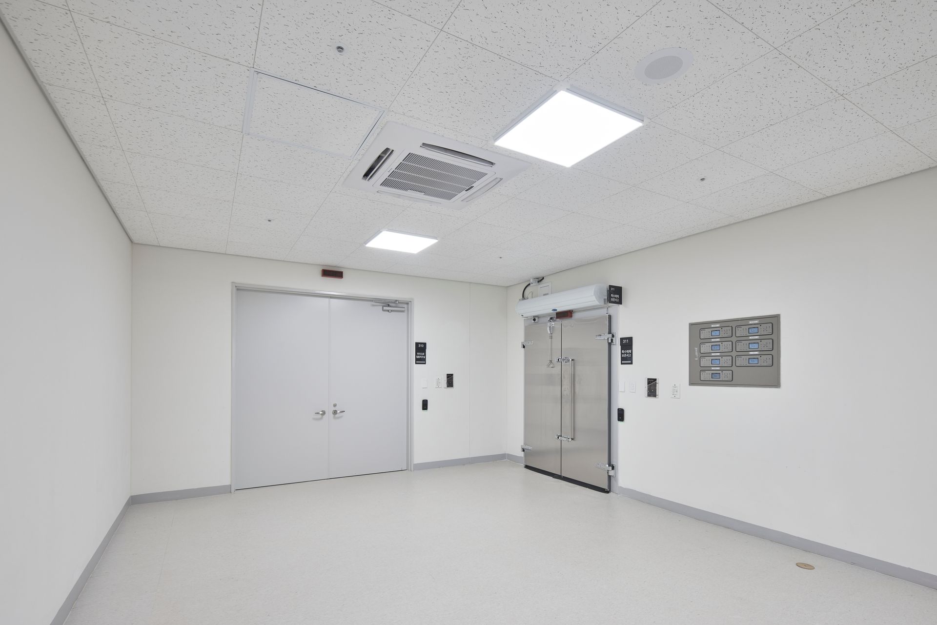 Pièce blanche vide avec double porte, panneau mural et grilles d'aération au plafond, éclairée par des néons.