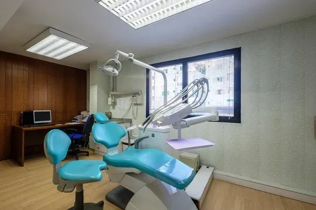 Consultorio dental con una silla color turquesa, un taburete de dentista, un escritorio de madera con una computadora
