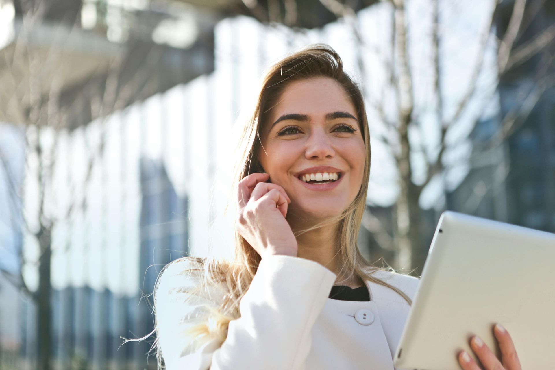 Una persona sonriente con una chaqueta blanca al aire libre, sosteniendo una tableta y hablando por teléfono celular.