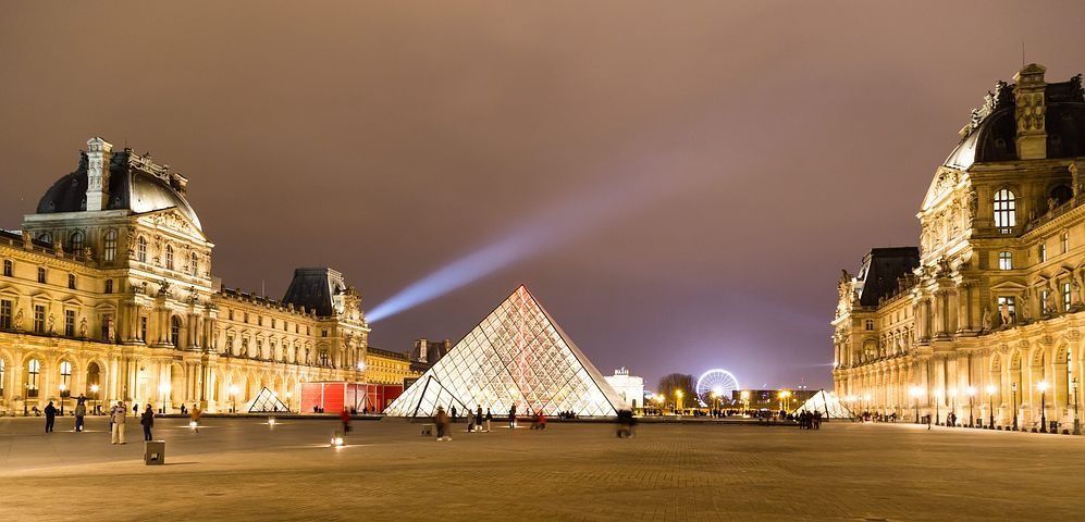 Vue nocturne du musée du Louvre à Paris, avec la pyramide de verre illuminée, entourée de bâtiments et du ciel illuminés.