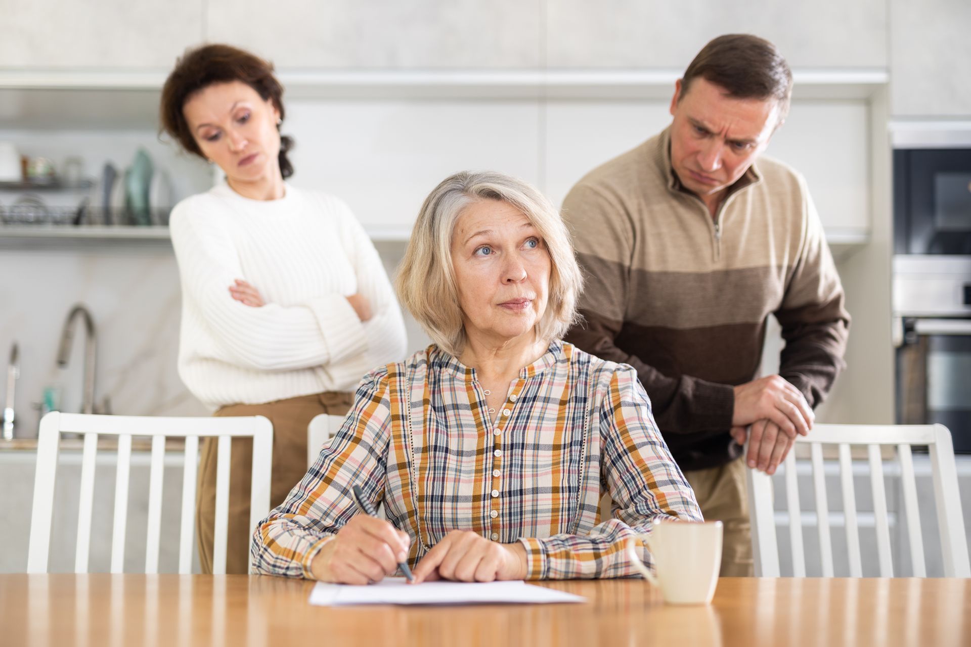 Eine ältere Frau unterschreibt an einem Tisch Dokumente; erwachsene Kinder schauen in einer Küche mit besorgten Gesichtern zu