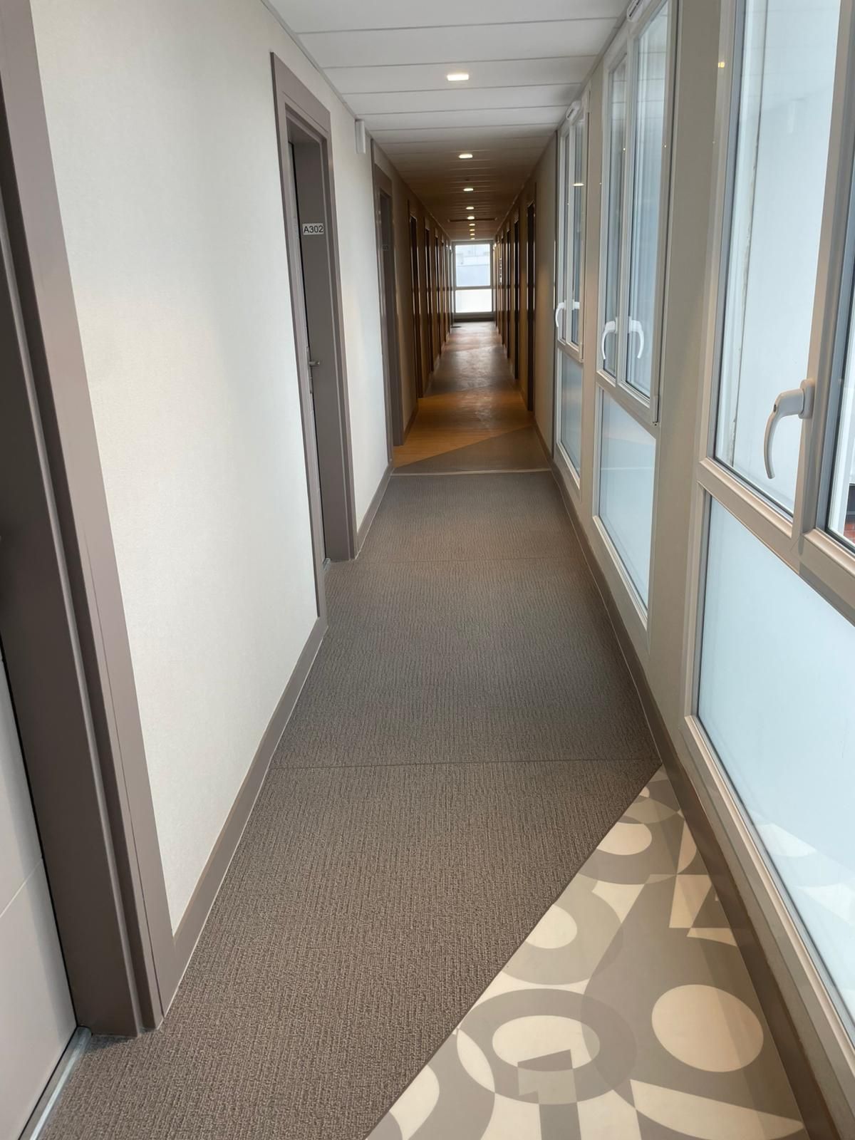 Couloir de bureau lumineux avec fenêtres à droite et moquette au sol menant à une porte au fond.