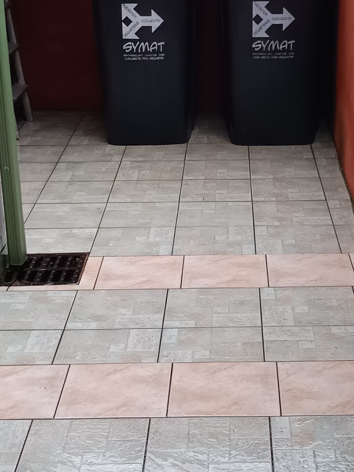 Couloir carrelé avec deux poubelles noires au fond et une bonde de sol sur la gauche.