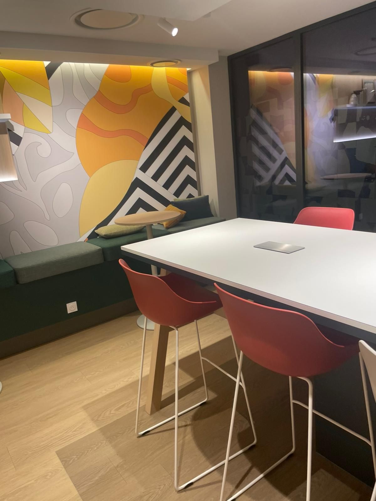 Salle de réunion moderne avec table blanche, chaises rouges, banc vert et fresque murale géométrique colorée