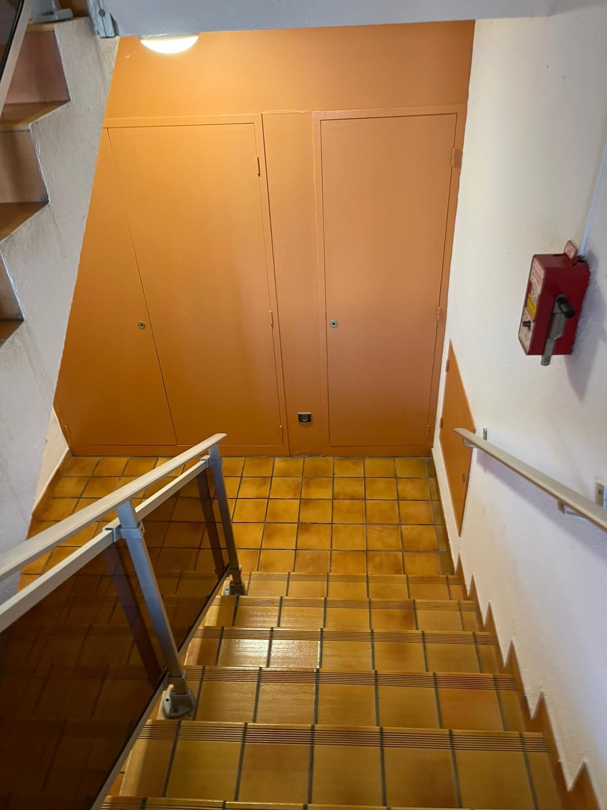 Escalier étroit avec sol et panneaux muraux carrelés orange, murs blancs, rampes et armoire à extincteurs rouge.
