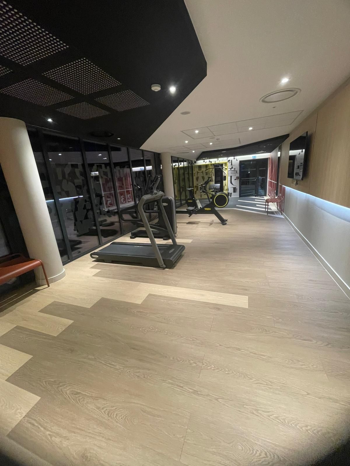 Intérieur de salle de sport moderne avec tapis de course, appareils de musculation et parquet sous un éclairage lumineux au plafond.