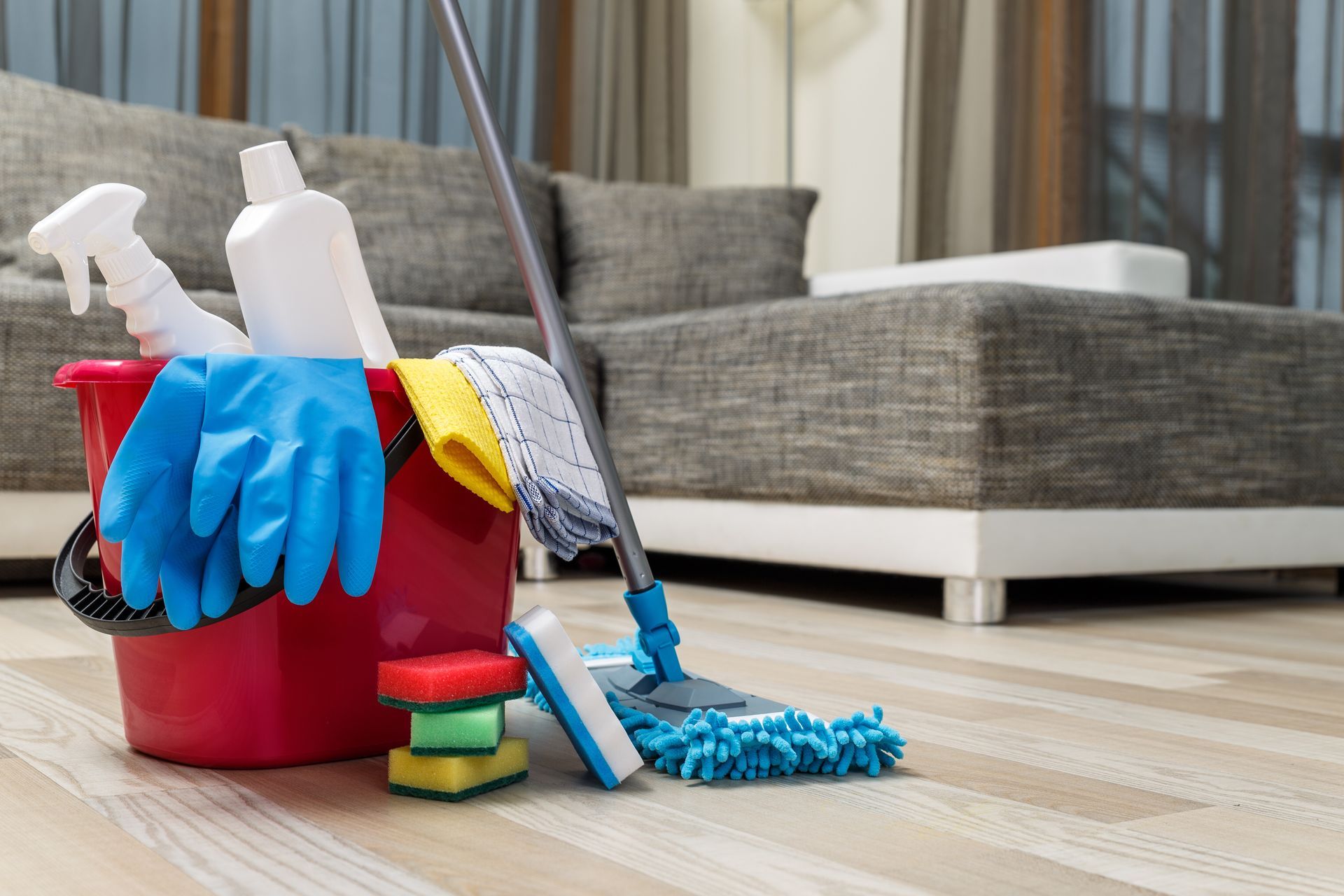 Des produits de nettoyage dans un seau rouge à côté d'une serpillière et d'un plumeau sur un parquet dans un salon