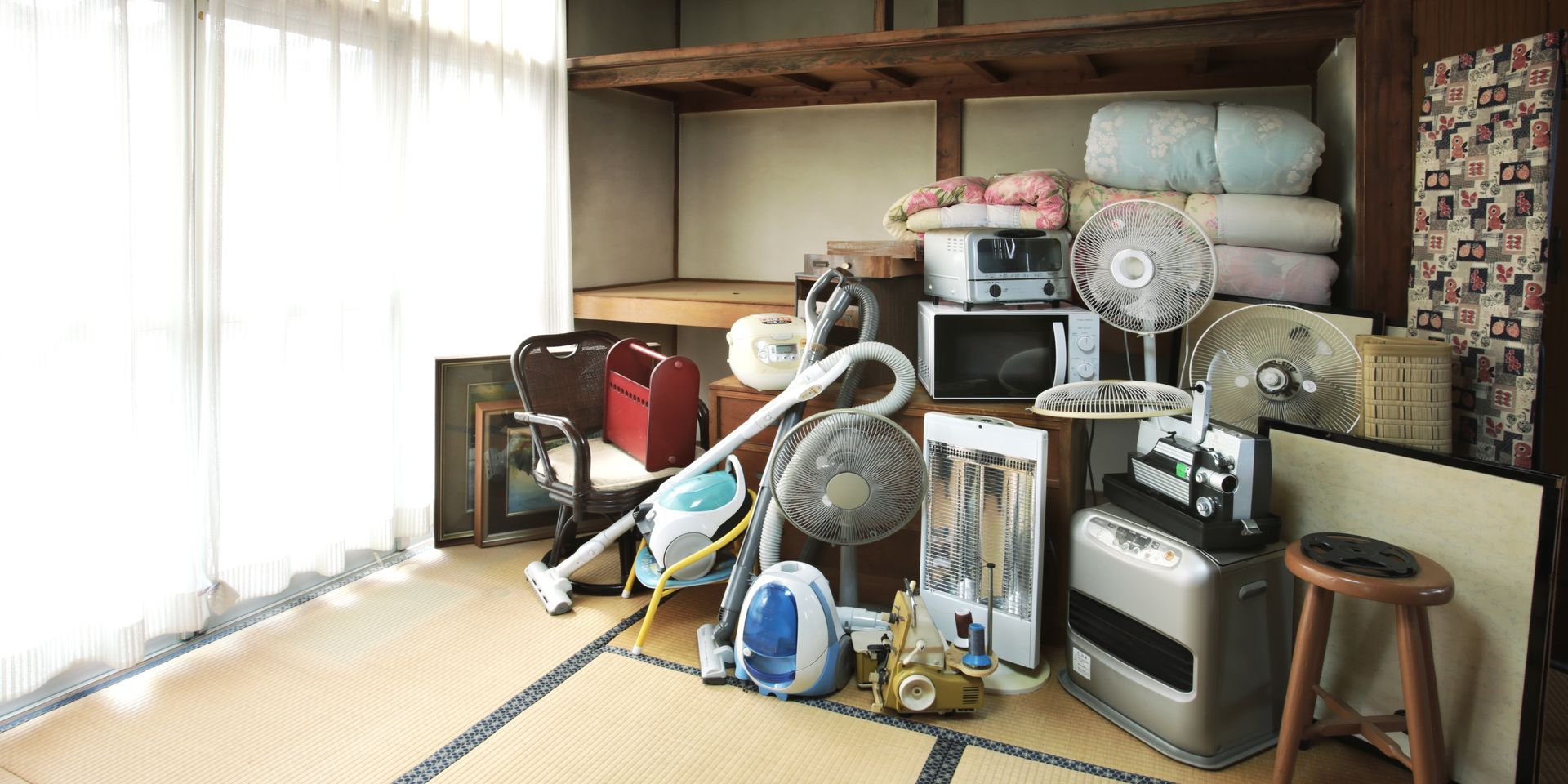 Pièce encombrée d'appareils électroménagers empilés, de cartons et d'un ventilateur près d'une fenêtre lumineuse