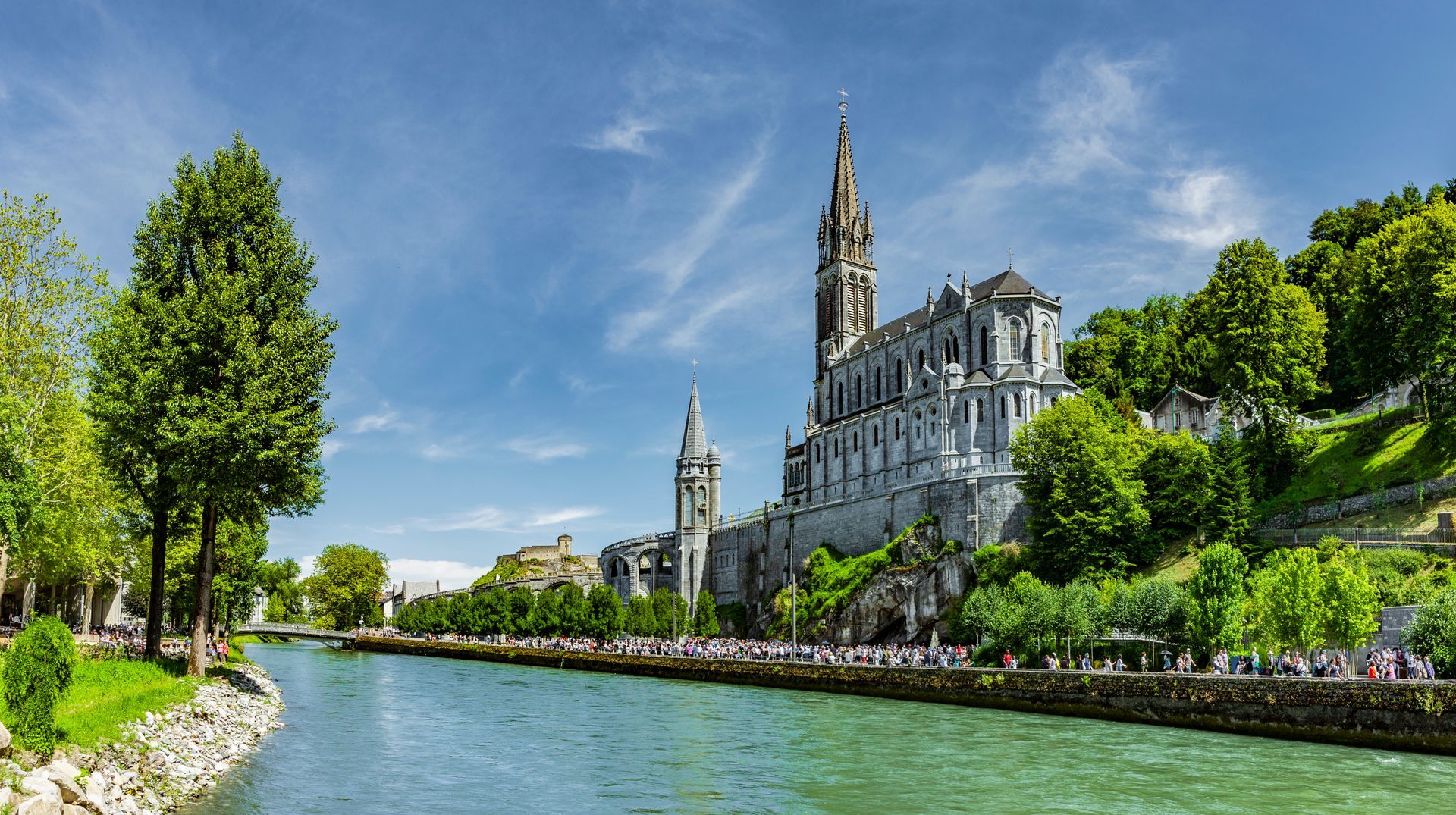Une rivière borde une cathédrale blanche et son clocher, sur une colline verdoyante et ensoleillée, sous un ciel bleu.