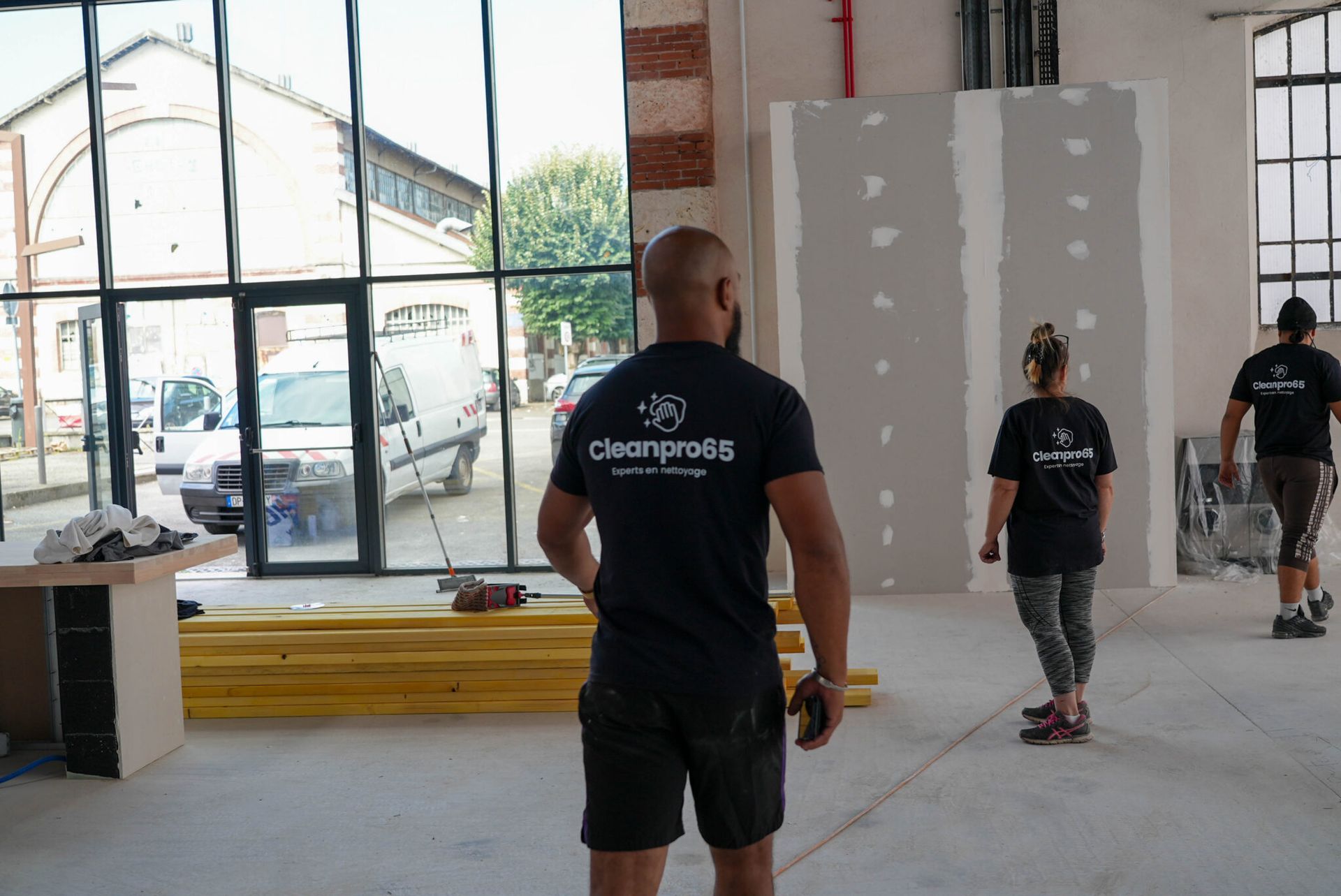 Des personnes se tiennent debout dans une pièce en travaux, entourées de plaques de plâtre et d'outils, comme si elles installaient ou déplaçaient du matériel.