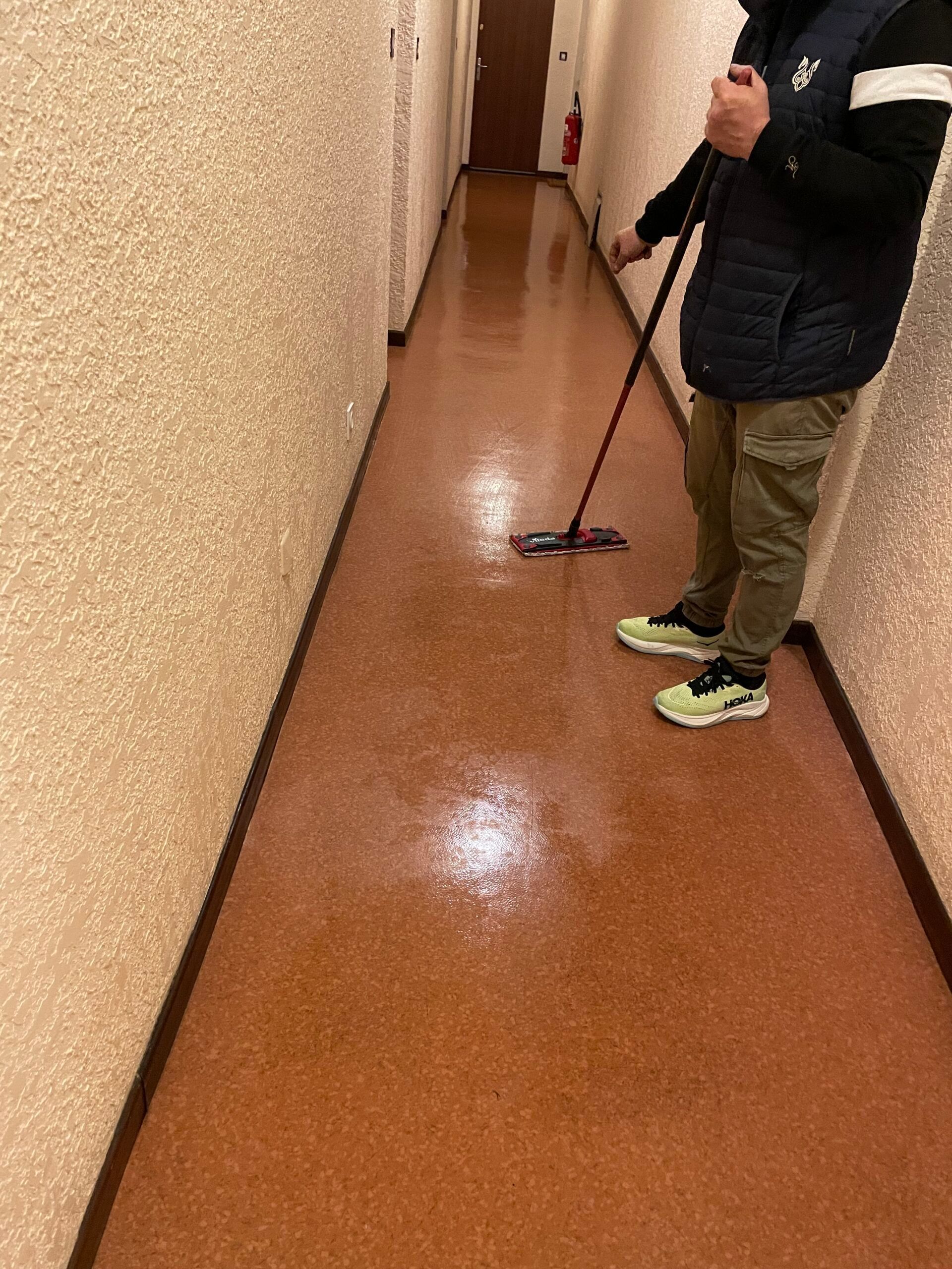 Une personne passe la serpillière sur le sol marron brillant d'un couloir, à côté d'un mur beige.