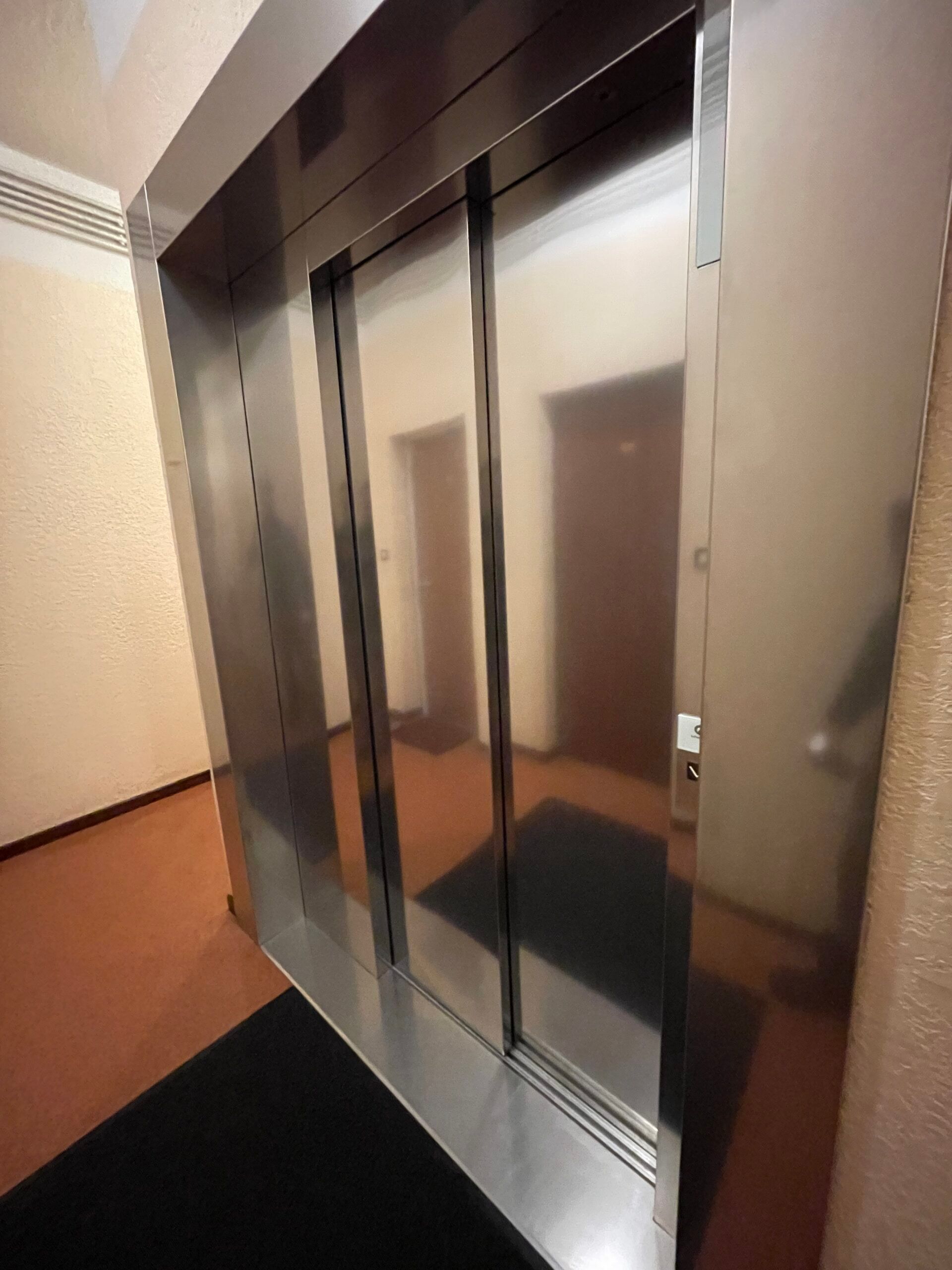 Portes d'ascenseur en acier inoxydable dans un couloir aux murs beiges et au tapis de sol foncé.