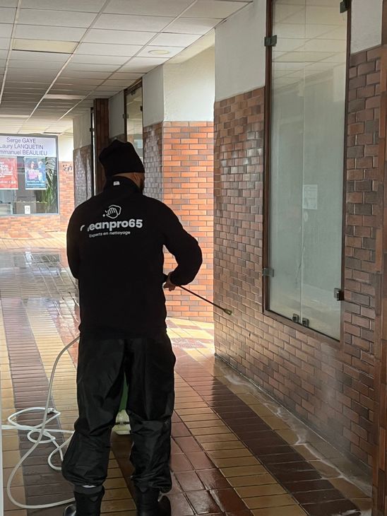 Une personne portant un sweat à capuche noir nettoie au nettoyeur haute pression un chemin en briques mouillées sous un couloir couvert d'un bâtiment.