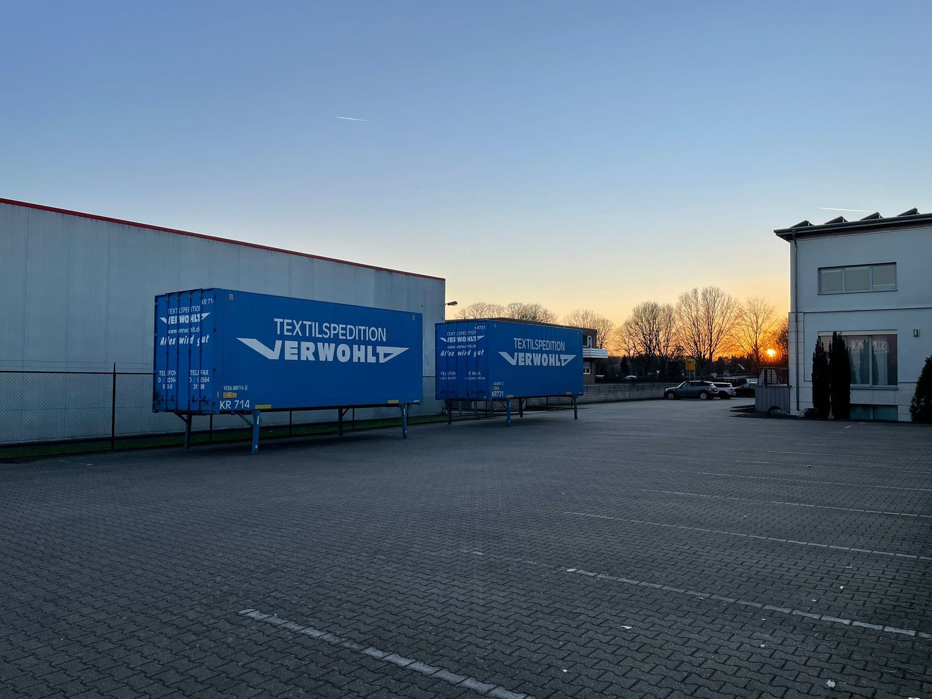 Zwei blaue Schiffscontainer stehen auf einem Parkplatz vor einem Gebäude.