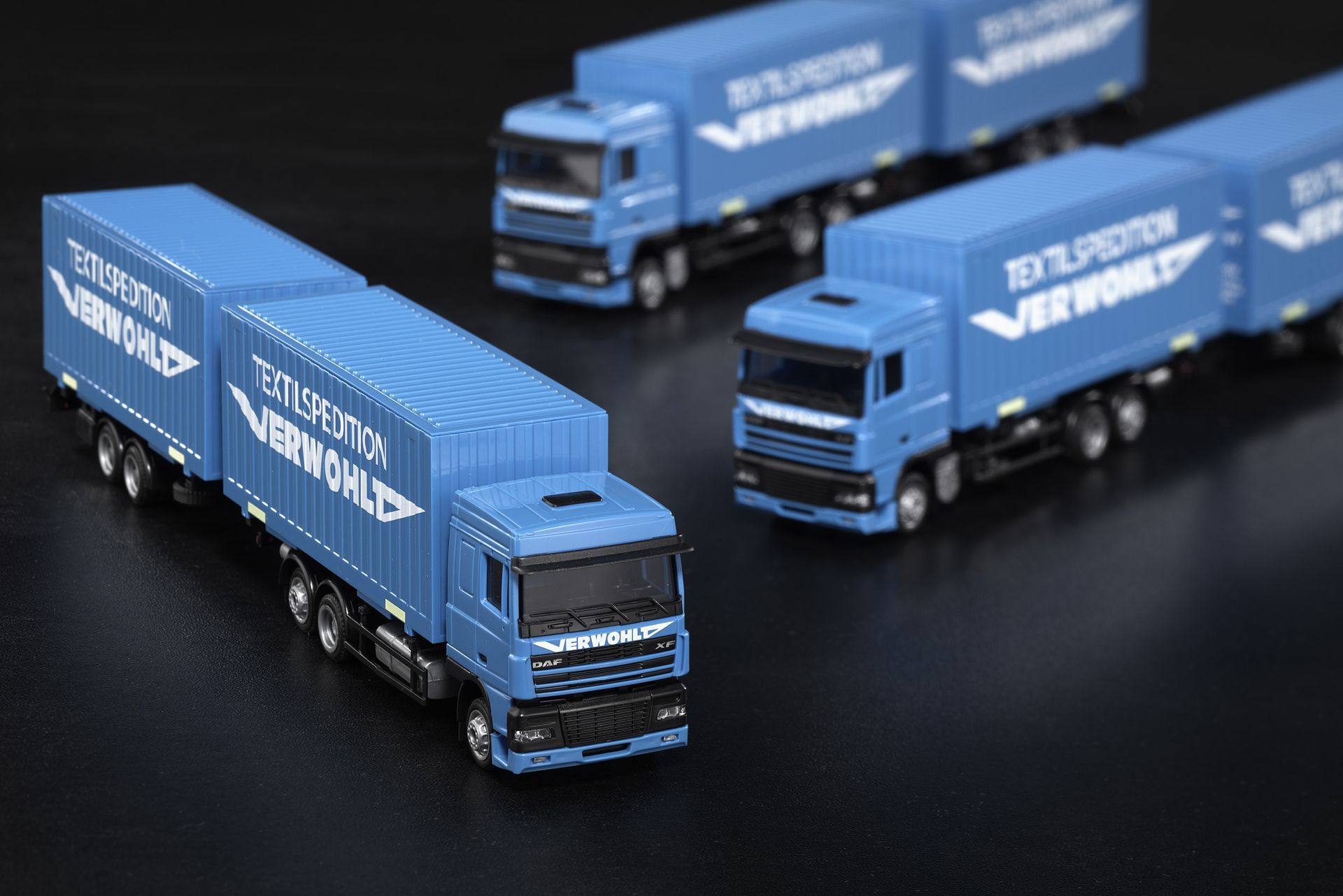 Drei blaue Spielzeuglastwagen stehen aufgereiht auf einer schwarzen Fläche.