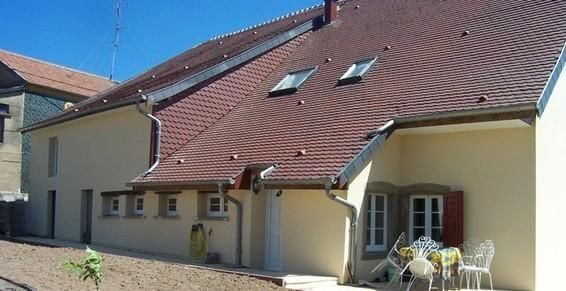 Maison beige au toit de tuiles rouges, dotée de deux puits de lumière et de fenêtres à cadre blanc ; extérieur ensoleillé.