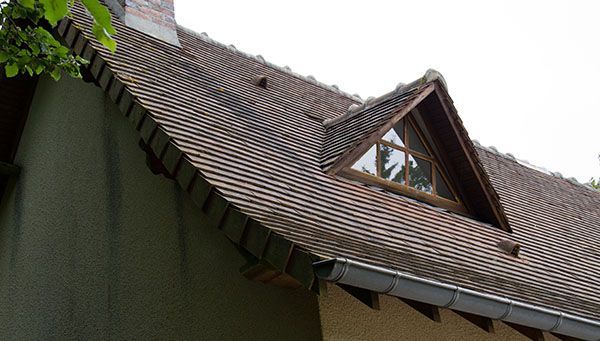 Toit en tuiles brunes avec une lucarne triangulaire, boiseries grises et cheminée en briques.