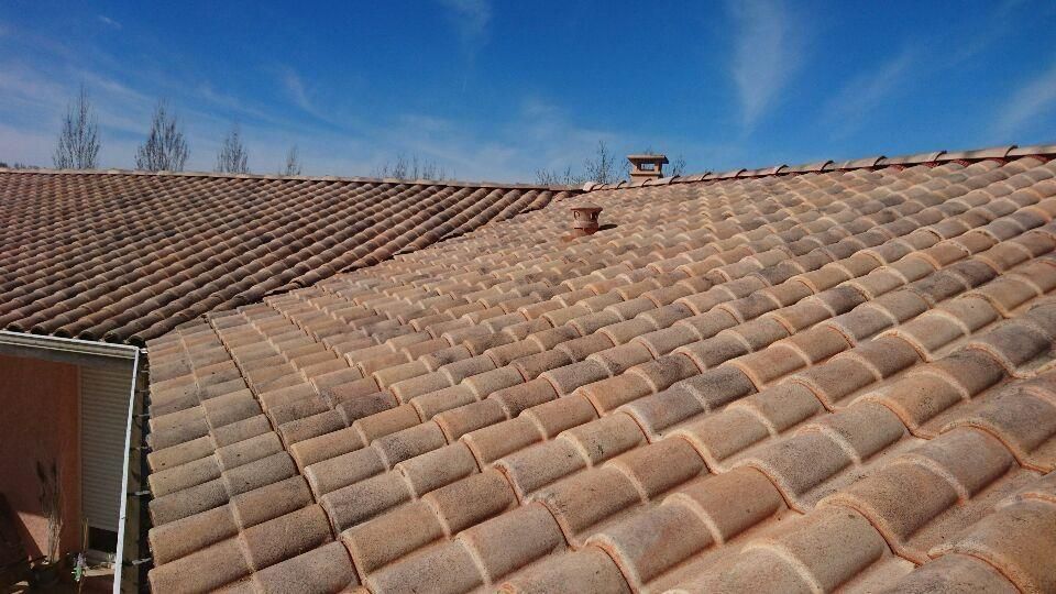 Toit de tuiles en terre cuite sous un ciel bleu limpide, avec une petite cheminée et des arbres au loin.