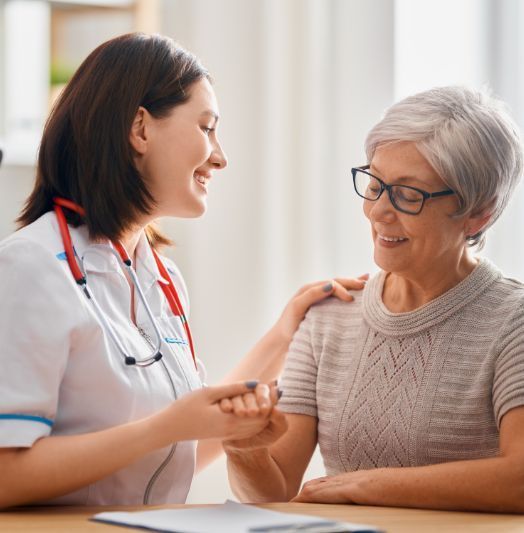 Eine Krankenschwester spricht mit einer älteren Frau, die an einem Tisch sitzt.
