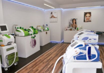 Interior de una clínica con diversos dispositivos médicos. Paredes blancas, suelo de madera y carteles promocionales.