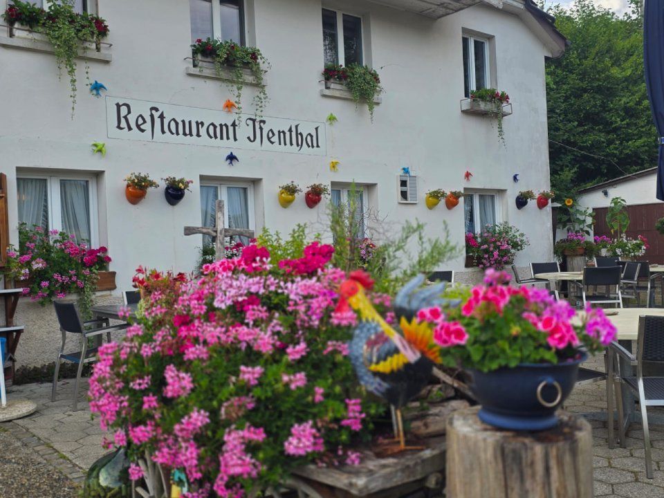 Blumenreiche Restaurantterrasse mit Hängeampeln und leuchtend rosa Blüten vor einem weissen Gebäude