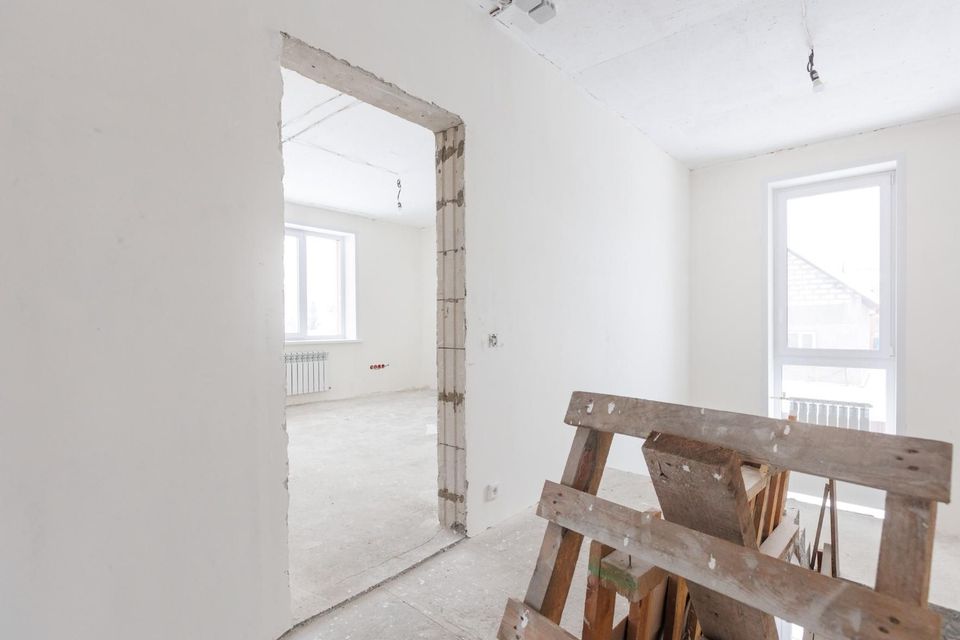 Habitación luminosa sin terminar con paredes blancas, puerta y tablas de madera apiladas en primer plano.