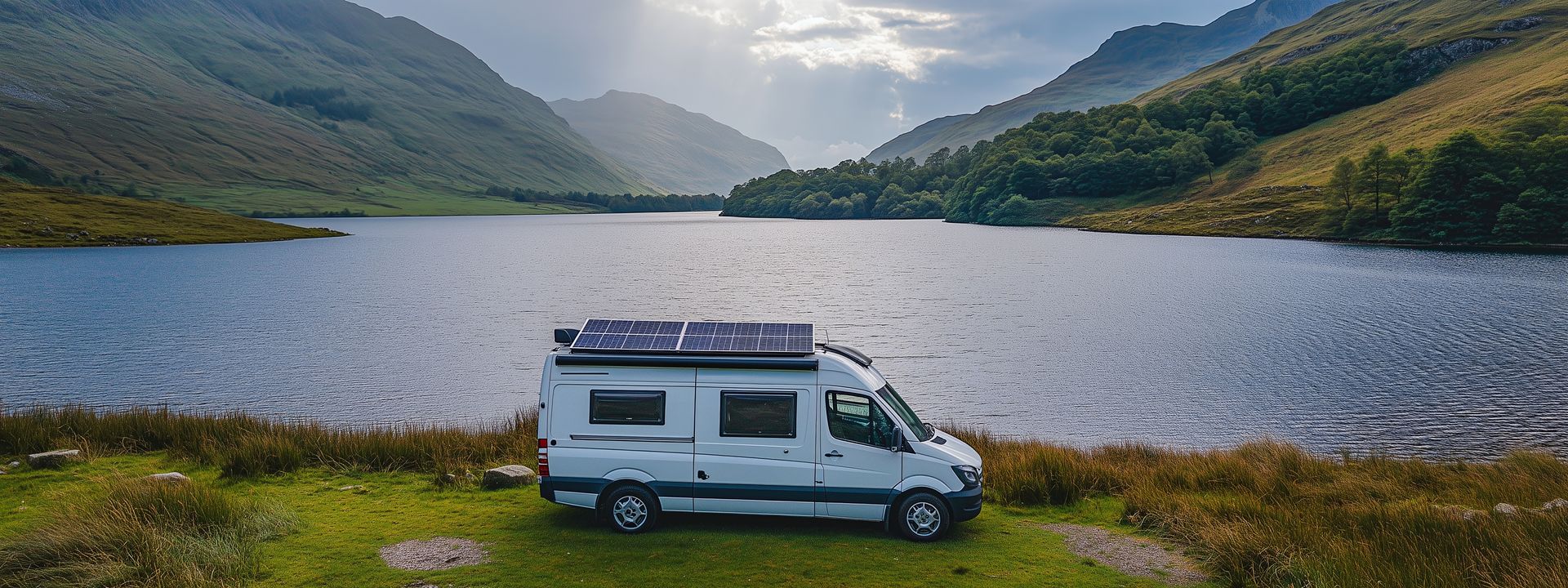 Un camping-car blanc garé sur la rive herbeuse d'un lac sombre, entouré de montagnes verdoyantes vallonnées sous un ciel nuageux.