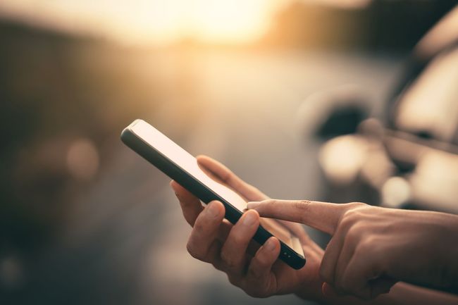 Personne utilisant un smartphone en extérieur, lumière du soleil en arrière-plan, voiture floue.